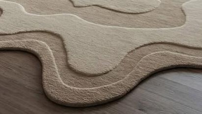 Pu&ograve; includere: Un tappeto beige e marrone chiaro con un design topografico a strati. Il tappeto presenta un motivo ondulato e organico con diverse tonalit&agrave; di beige e marrone chiaro, creando un effetto strutturato e tridimensionale. Il design del tappeto assomiglia a un paesaggio.