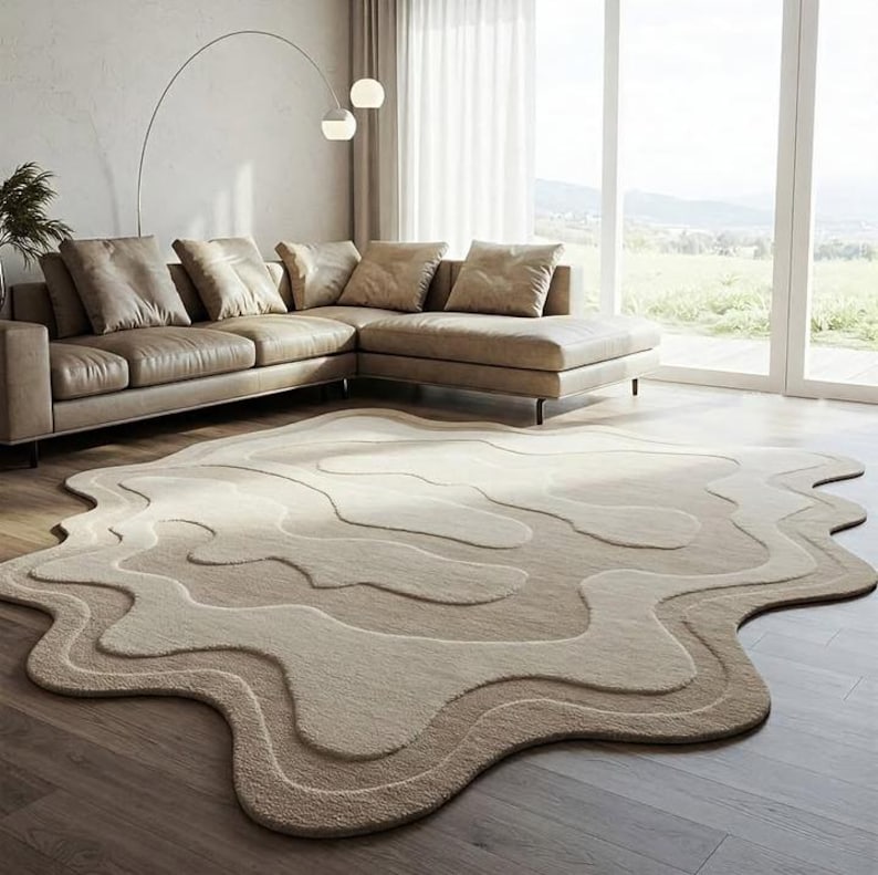 Pu&ograve; includere: Un tappeto beige con una forma organica unica, caratterizzato da motivi a strati e fluidi in varie tonalit&agrave; di beige e crema. Il tappeto &egrave; posizionato su un pavimento in legno in un soggiorno, completando i toni neutri del divano e dell'arredamento.