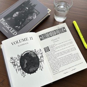 Op de afbeelding: Een open boek, "VOLUME II FLASHBACK", met zwart-wit illustraties en tekst. Een gesloten boek, "MANACLED", en een glas water zijn ook zichtbaar. Een gele markeerstift ligt op het houten oppervlak.