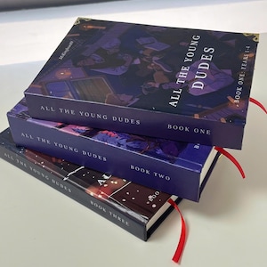 May include: Three hardcover books stacked, featuring the title "ALL THE YOUNG DUDES." The covers are dark blue with illustrations and text. Red ribbon bookmarks protrude from the books. The books are labeled "BOOK ONE," "BOOK TWO," and "BOOK THREE."