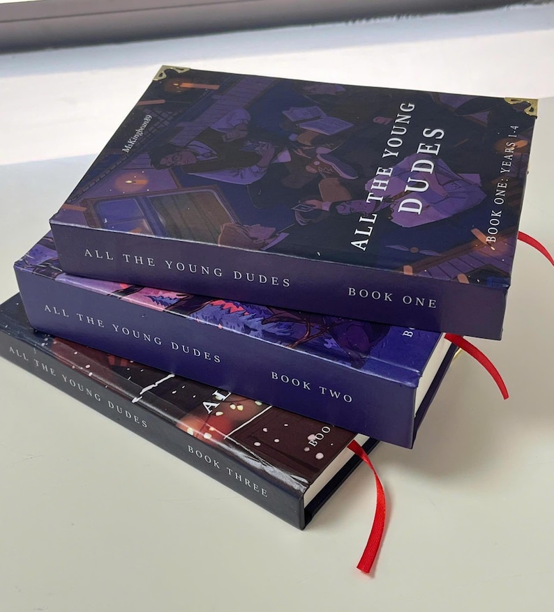 May include: Three hardcover books stacked, featuring the title "ALL THE YOUNG DUDES." The covers are dark blue with illustrations and text. Red ribbon bookmarks protrude from the books. The books are labeled "BOOK ONE," "BOOK TWO," and "BOOK THREE."