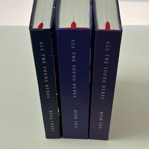 May include: Three hardcover books titled "ALL THE YOUNG DUDES" in varying shades of blue. Each book has a red bookmark ribbon. The spines display "BOOK ONE", "BOOK TWO", and "BOOK THREE".