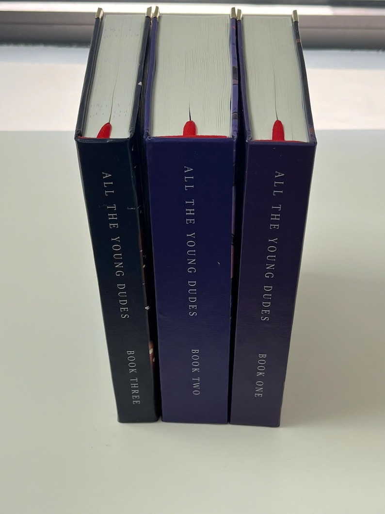 May include: Three hardcover books titled "ALL THE YOUNG DUDES" in varying shades of blue. Each book has a red bookmark ribbon. The spines display "BOOK ONE", "BOOK TWO", and "BOOK THREE".