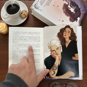Könnte beinhalten: Ein offenes Buch mit Illustrationen von Charakteren, eine Tasse Kaffee, ein Keks und eine brennende Kerze. Das Buch zeigt eine Zeichnung eines Mannes mit einem Totenkopf-Tattoo und einer Frau. Eine Brille und ein Stift liegen ebenfalls auf dem Tisch.