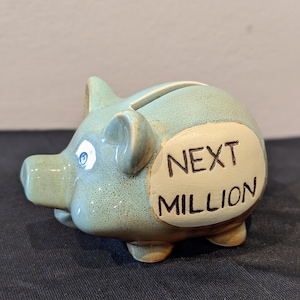 Könnte beinhalten: Ein hellblaues Keramik-Sparschwein mit der Aufschrift "NEXT MILLION" auf einem cremefarbenen Oval. Das Sparschwein hat einen Schlitz zum Einwerfen von Geld und ist eine unterhaltsame Art zu sparen.