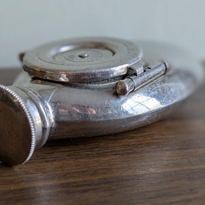 Puede incluir: Un frasco antiguo de color plateado con tapa abisagrada y tapón de rosca. El frasco tiene forma redondeada y está sobre una superficie de madera. La tapa es circular con un diseño decorativo.