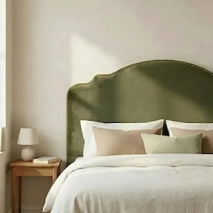 May include: An olive green velvet headboard with a curved top edge and brass nailhead trim. The headboard is positioned behind a bed dressed in white linens, featuring two beige rectangular pillows and one smaller light green lumbar pillow.