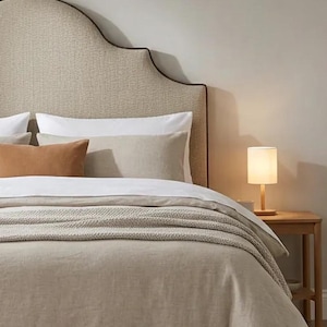 May include: A bed featuring a beige upholstered headboard with a dark brown trim, white and beige bedding, a solid rust-coloured rectangular pillow, a textured beige throw blanket, and a small wooden table lamp with a white cylindrical shade.