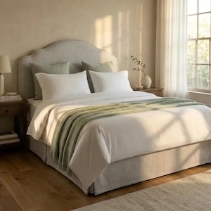 May include: A bed featuring a curved, striped headboard and matching bed skirt, dressed in white bedding with two white pillows, two sage green accent pillows, and a sage green throw blanket draped across the foot of the mattress.