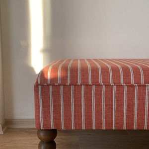 May include: A rectangular bench upholstered in a fabric featuring vertical stripes in shades of terracotta orange and off-white. The bench is supported by a dark brown wooden leg with a stacked spherical, ball-shaped design at the base.
