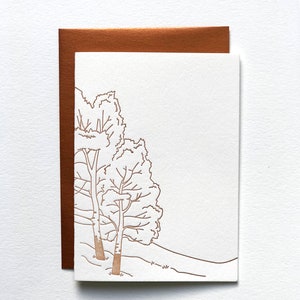 May include: A blank greeting card with a copper-coloured envelope. The card features a detailed line drawing of two trees on a hillside. The trees and the hillside are in a copper colour, and the background is white.