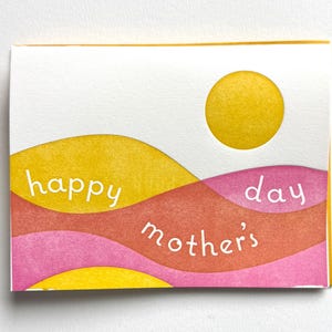 May include: A greeting card with a yellow sun and layered, wavy bands of yellow, orange, and pink. The words "happy mother's day" are written across the card in white. The card is rectangular with a white background and yellow border.