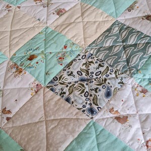 May include: A patchwork quilt featuring a repeating pattern of squares in mint green, cream, and white. The fabric displays illustrations of small woodland animals, floral motifs, paisley designs, and the text Once upon a time in script.