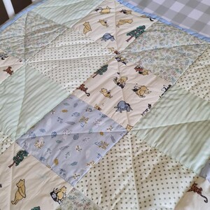 May include: A quilted baby blanket featuring a patchwork design with alternating squares of Winnie the Pooh character prints, light green stripes, sage green polka dots, and floral patterns in soft pastel shades of cream, blue, and green.