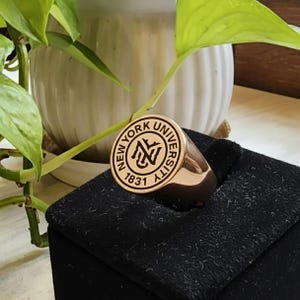 Może przedstawiać: Złoty sygnet z napisem "NEW YORK UNIVERSITY" i rokiem "1831". Na środku znajduje się stylizowane logo "NYU". Pierścień jest prezentowany na czarnej powierzchni.