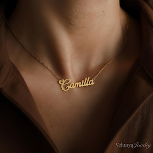 Pode incluir: Um colar dourado com o nome "Camilla" em fonte cursiva. O colar é exibido no pescoço de uma pessoa, com uma camisa marrom ao fundo. A joia é da Velunya Jewelry.