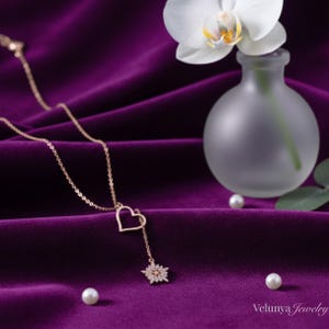 Op de afbeelding: Een goudkleurige ketting met een hartvormige hanger en een hangend bloemmotief, tegen een dieppaarse fluwelen achtergrond. De ketting wordt getoond met een kleine vaas met een witte orchidee en verspreide parels.