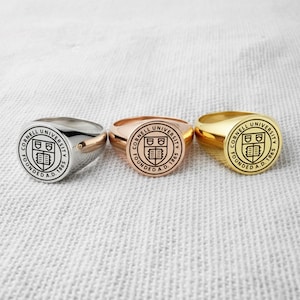 Könnte beinhalten: Drei Siegelringe in Silber, Roségold und Gold. Jeder Ring zeigt das Siegel der Cornell University mit dem Text "CORNELL UNIVERSITY FOUNDED A.D. 1865" in einem kreisförmigen Design. Die Ringe werden auf einer weißen, strukturierten Oberfläche präsentiert.