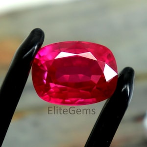 Puede incluir: Un vibrante rubí de talla cojín sostenido por pinzas negras. El rubí muestra un rico tono rojo saturado con facetas que capturan la luz. El texto "EliteGems" es visible en la imagen.