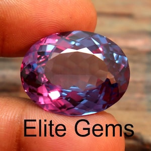 Può includere: Una gemma ovale sfaccettata, tenuta tra le dita. La gemma mostra un cambio di colore dal rosa al blu, con una superficie lucida. Il testo "Elite Gems" è visibile in basso.
