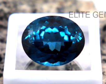 Puede incluir: Una gema ovalada facetada de color verde azulado oscuro, engastada en una vitrina blanca. La gema tiene un corte brillante, reflejando la luz con una apariencia vibrante y multifacética. El texto "ELITE GEMS" es visible.