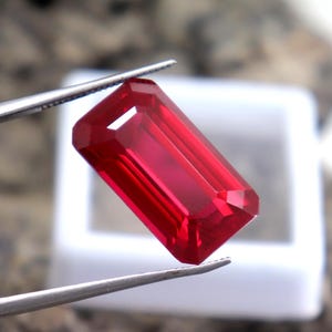 Op de afbeelding: Een gefacetteerde, rechthoekige, dieprode edelsteen, vastgehouden door een zilveren pincet. De edelsteen heeft een helder, gepolijst oppervlak en staat tegen een vage achtergrond, wat op een sieraad wijst.