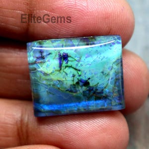 May include: A square gemstone with a vibrant display of blues, greens, and hints of black. The stone has a polished surface and a unique, natural pattern. The text "EliteGems" is visible.