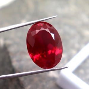 Puede incluir: Una gema de rubí facetada, de forma ovalada, sujeta con pinzas. El rubí es de un color rojo intenso y vibrante, con múltiples facetas que captan la luz. El fondo es borroso, de tono neutro, con un cuadrado blanco.