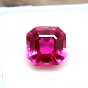 May include: A faceted, rectangular, hot pink gemstone set in a white plastic case. The gem has a brilliant cut, reflecting light. The logo "EliteGems" is visible in the upper left corner. The background is a textured, brown wooden surface.