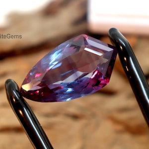 May include: A colour-shifting, faceted gemstone, transitioning from purple to blue. Held by black tweezers against a blurred wooden background. The text "EliteGems" is visible.