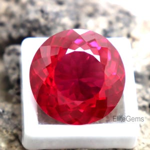 Könnte beinhalten: Ein facettierter, runder, tiefroter Edelstein, möglicherweise ein Rubin, in einer weißen Präsentationsbox. Der Edelstein hat eine hohe Klarheit und Brillanz und reflektiert das Licht. Der Hintergrund ist eine verschwommene, strukturierte Oberfläche.
