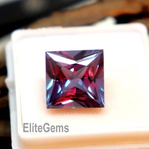 Puede incluir: Una gema de alejandrita de corte cuadrado, presentada en una caja blanca. La gema exhibe un efecto de cambio de color, con tonos morados y turquesas. El texto "EliteGems" es visible. El fondo es un trozo de madera.