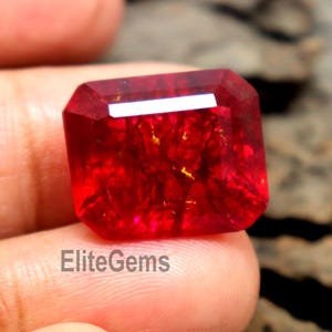 Puede incluir: Una gema roja rectangular facetada con un corte brillante. La gema se sostiene entre los dedos, mostrando su color vibrante y estructura interna. El texto "EliteGems" es visible.