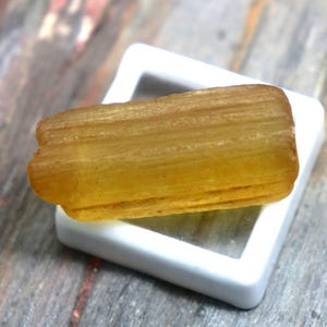 May include: A golden-yellow, rectangular gemstone rests inside a white square display. The gemstone has a textured surface and is displayed on a wooden surface with gray and brown tones. The gemstone is a natural mineral.