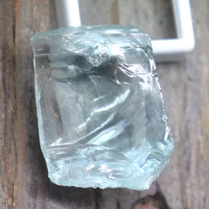 Könnte beinhalten: Ein hellblauer, grob geschnittener Edelstein mit kristallinem Aussehen. Der Stein ist vor dem Hintergrund von verwittertem Holz und einem weißen, quadratischen Rahmen platziert.