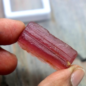 Könnte beinhalten: Ein roher, rechteckiger, rosa Turmalin-Edelstein. Der Kristall wird zwischen den Fingern gehalten und zeigt seine natürliche, unpolierte Textur und Farbvariationen. Der Stein hat eine durchscheinende Qualität mit sichtbaren inneren Linien.