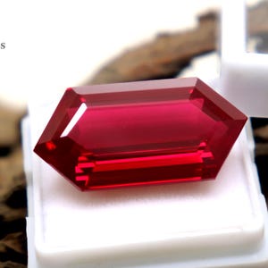 May include: A faceted, rectangular-cut, deep red gemstone rests in a white display case. The gem has a hexagonal shape with multiple facets, reflecting light. The background is a textured, brown surface. The text "EliteGems" is visible.