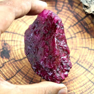 May include: A rough, uncut ruby gemstone with a deep red hue. The ruby is held in a hand, showcasing its irregular shape and textured surface. The background is a wooden surface with a natural grain pattern.