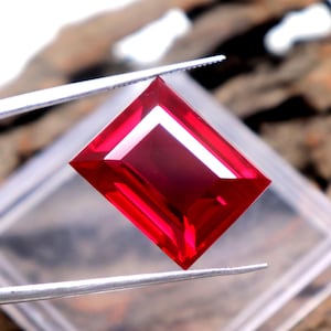 Op de afbeelding: Een levendige, rechthoekig geslepen rode edelsteen vastgehouden door een zilverkleurige metalen pincet. De steen heeft scherpe, heldere facetten die het licht reflecteren, wat zorgt voor een diepe karmozijnrode tint met heldere accenten.