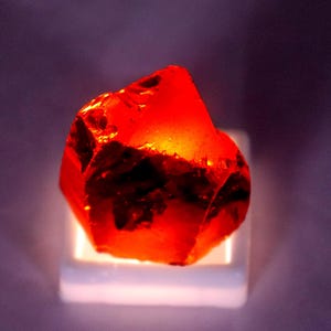 Puede incluir: Un espécimen de cristal en bruto de color naranja rojizo translúcido con bordes irregulares y dentados, y una apariencia facetada. El cristal está iluminado desde abajo por una base de luz cuadrada blanca que proyecta un brillo intenso a través.