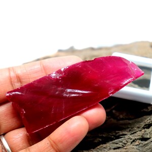 Op de afbeelding: Een levendige, ruwe robijn edelsteen met een dieprode kleur. De ruw gesneden steen wordt in een hand gehouden, waardoor de natuurlijke textuur en kleur zichtbaar worden. Een kleine witte vitrine is zichtbaar op de achtergrond, wat duidt op een verzamelobject.