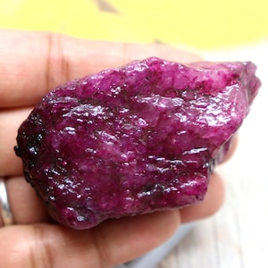 Puede incluir: Una piedra preciosa de rubí en bruto y sin pulir con un tono magenta y púrpura intenso. La piedra presenta una superficie rugosa y texturizada con facetas cristalinas naturales e inclusiones oscuras en toda su forma irregular y alargada.