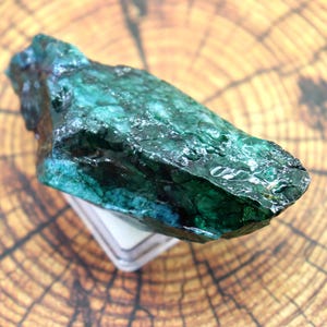 Puede incluir: Un espécimen mineral en bruto y sin pulir que presenta tonos profundos de verde bosque y azul verdoso con una textura superficial cristalina y brillante. La piedra tiene una forma irregular y alargada con bordes dentados e inclusiones opacas.