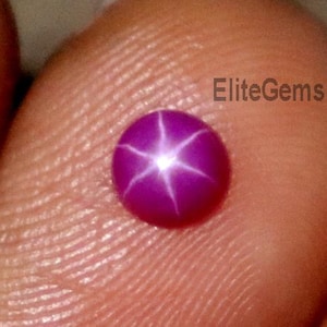May include: A small, round, purplish-red gemstone with a six-pointed star pattern. The gem is held between fingers, with the text "EliteGems" visible. The star effect is created by light reflecting within the stone.