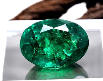 Könnte beinhalten: Ein lebendiger, oval geschliffener Smaragd in einer weißen Box. Der Edelstein hat eine tiefe grüne Farbe und reflektiert das Licht, wodurch seine Facetten zur Geltung kommen. Die Box liegt auf einer strukturierten, braunen Holzoberfläche. Der Text "EliteGems" ist sichtbar.