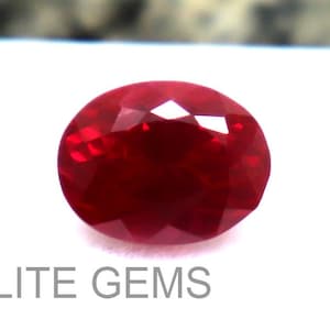 Pode incluir: Uma pedra preciosa de rubi facetada em forma oval. A gema vermelha escura é exibida em uma superfície branca, com o texto "ELITE GEMS" visível abaixo. O corte da gema permite a reflexão da luz.