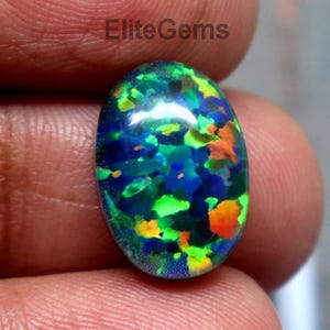 Puede incluir: Una piedra preciosa de ópalo ovalada con una vibrante exhibición de colores. El ópalo presenta una base oscura con destellos de azul, verde y naranja. La gema se sostiene entre los dedos, mostrando su superficie pulida y belleza natural.