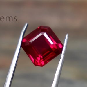 Puede incluir: Un rubí tallado en forma cuadrada, sujeto con pinzas. La gema, de un rojo intenso y vibrante, presenta facetas que capturan la luz. El fondo está borroso, resaltando la joya. El texto "EliteGems" es visible en la esquina superior izquierda.