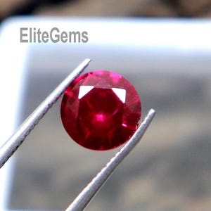 May include: A faceted, round, deep red gemstone is held by silver tweezers. The gem is displayed against a white background inside a square container. The text "EliteGems" is visible in the background.