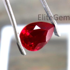 May include: A deep red, pear-cut ruby gemstone held by tweezers. The gem's facets reflect light. The image includes the text "Elite Gems". The ruby is positioned above a white square. The gem is a vibrant, rich red colour.
